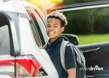 Student wearing a backpack stands beside an open passenger vehicle in an EverDriven alternative student transportation image.