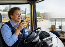 A female school bus driver in bus driver's seat speaking on radio with an inset of female with headphones looking at two monitors and buses in the background
