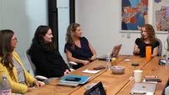 Four women participate in a council meeting around a wooden conference table, with one speaking while the others listen. Laptops, notebooks, and drinks sit on the table.