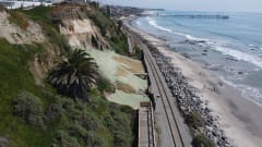 Coastal railroad tracks run between a steep bluff and the Pacific Ocean in San Clemente, California, with rock barriers protecting portions of the shoreline.