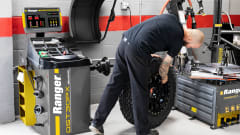 Automotive technician handling a large tire and wheel assembly beside a Ranger DST30PX wheel balancer and tire-changing equipment in a professional repair shop.