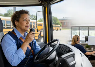 A female school bus driver in bus driver's seat speaking on radio with an inset of female with headphones looking at two monitors and buses in the background