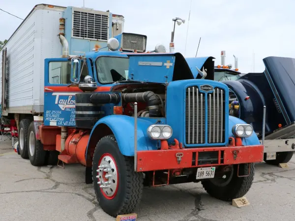 A blue, black, and red conventional truck from the 1950s with a trailer on display in a parking lot