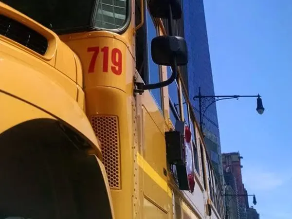 A yellow school bus is parked on a street in Lower Manhattan beneath One World Trade Center. The low-angle photo captures the towering skyscraper rising above the bus against a clear blue sky, contrasting student transportation with the iconic New York City skyline.