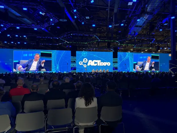 Big convention hall area with seating photo shot from back of room showing speaker on video screens