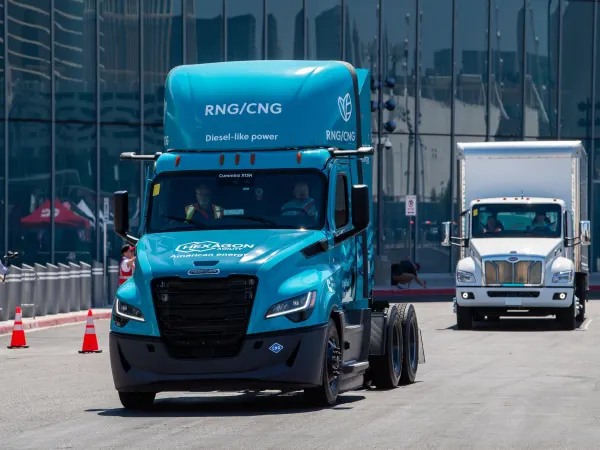 Blue RNG/CNG powered truck driving on closed course with medium duty white box truck behind it