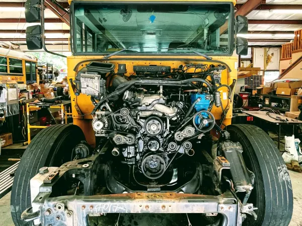 A yellow school bus sits inside a maintenance shop with its front bodywork removed, exposing the engine, belts, pulleys, and mechanical components. The image offers a detailed look at a bus undergoing major repair or maintenance.