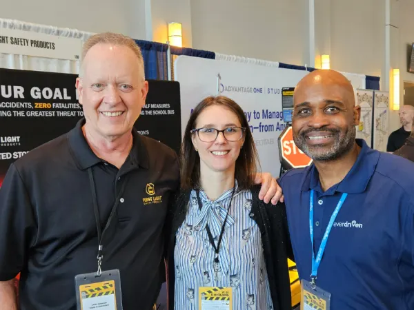 Three people smile together on the vendor expo floor, standing in front of exhibit booths and safety product displays.