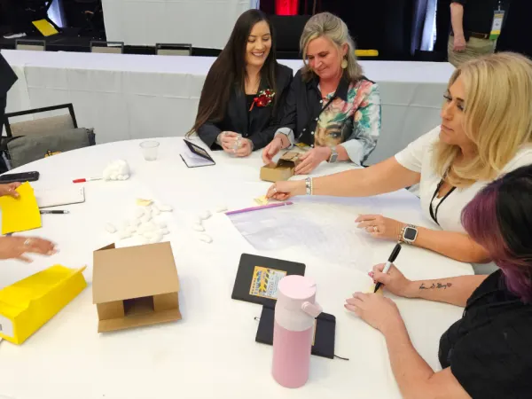 Small group of attendees participate in a hands-on workshop craft activity at a round table, using materials to assemble a paper school bus as part of a learning exercise.