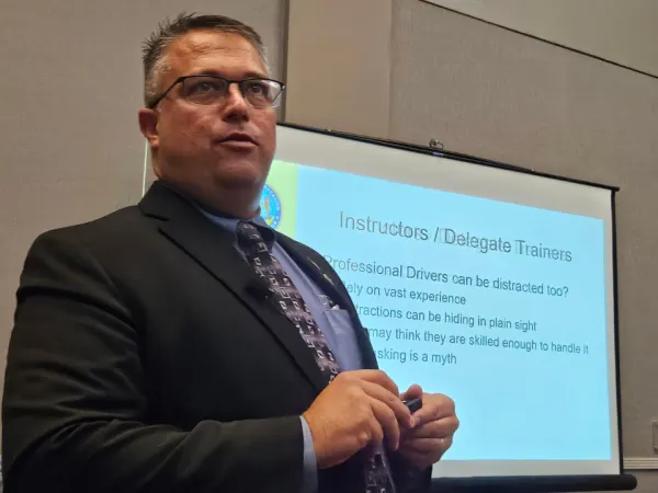 Presenter in a suit speaks during a breakout session, standing near a projection screen displaying training slides for instructors and delegate trainers.