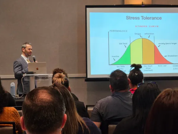 A speaker presents on stress tolerance and burnout, standing beside a projected graph illustrating performance levels and stress curves during an educational session.