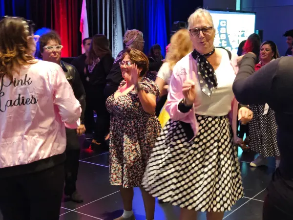 Conference attendees dressed in retro “Grease”-themed outfits dance together at the evening banquet, celebrating during the themed dinner event.