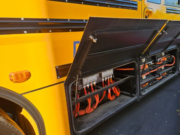 Close-up of an electric school bus with side compartments open, revealing battery and high-voltage components during an equipment demonstration.