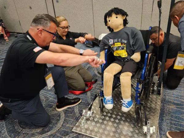 Participants practice wheelchair securement techniques using a training mannequin and tie-down equipment during a safety demonstration.