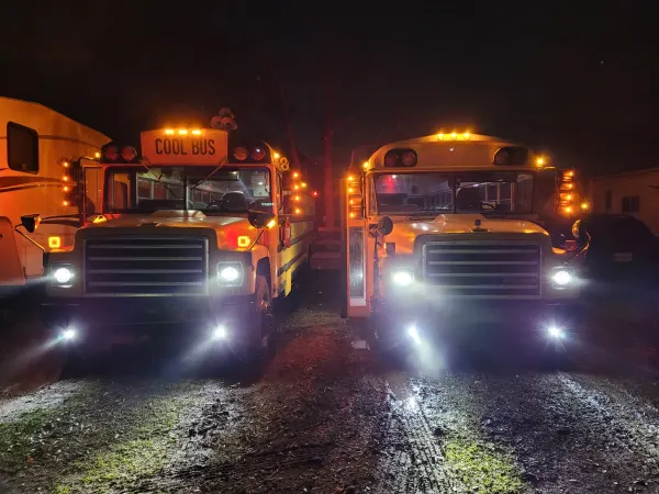 Two vintage school buses are parked side by side at night with headlights, marker lights, and warning lights illuminated. One bus displays a playful "COOL BUS" destination sign, creating a nostalgic scene with warm amber lighting.