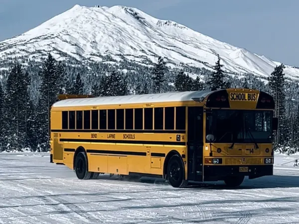 A yellow Bend-La Pine Schools school bus sits on a snow-covered lot with a towering snow-capped mountain in the background. Evergreen trees and winter conditions showcase the challenges of transporting students in alpine terrain.