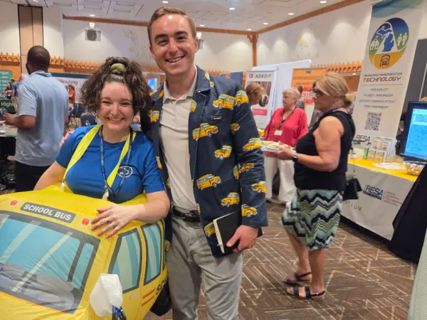 BusRight team members pose in school bus-themed outfits during the conference trade show, including an inflatable school bus costume and a jacket decorated with buses.