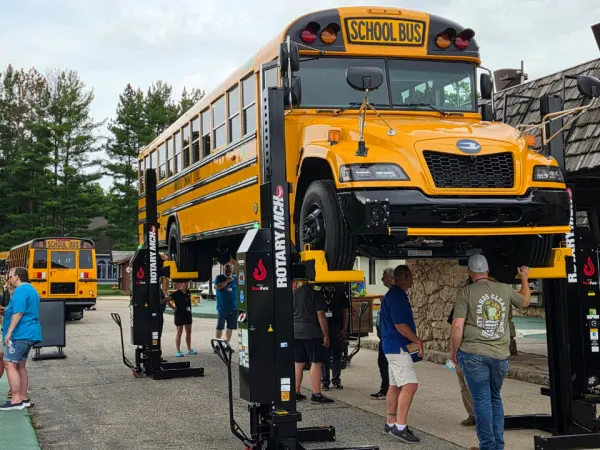 A new Blue Bird school bus is lifted on a heavy-duty Rotary vehicle hoist during an outdoor maintenance equipment demonstration as attendees gather nearby.