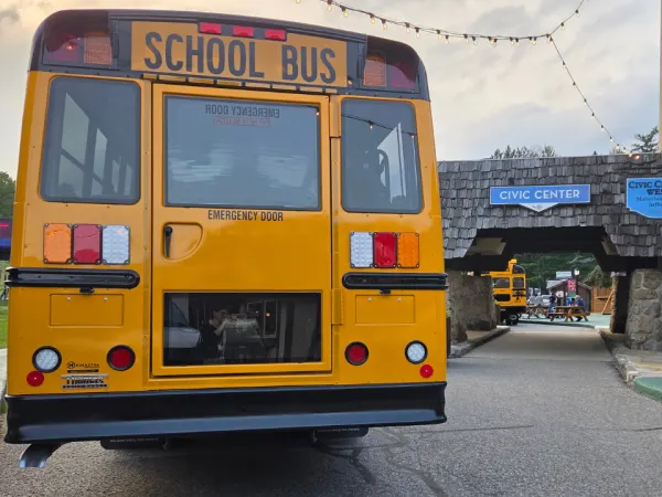 A yellow school bus is parked outside Boyne Mountain Resort near the Civic Center entrance, with string lights overhead and another school bus visible through the covered walkway in the background.