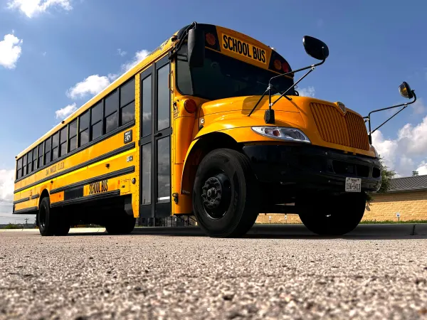 Low-angle view of a yellow International conventional school bus parked on pavement under a bright blue sky with scattered clouds. The bus is photographed from the front driver's side, highlighting the hood, crossing mirrors, warning lights, and full passenger body. The side reads "San Marcos CISD," and the image emphasizes the size and visibility of the school bus.