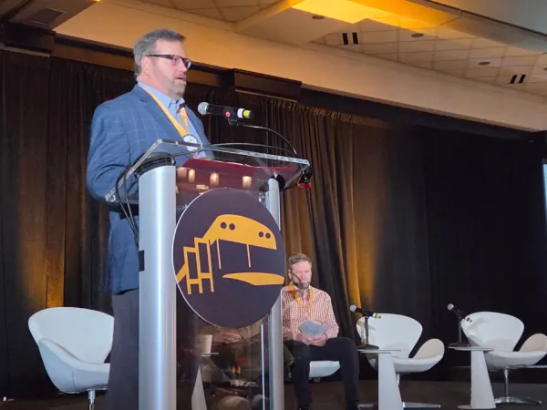 A speaker addresses NAPT ACTS 2026 attendees from the podium while panelists sit behind him and a transportation industry update slide appears on the screen.