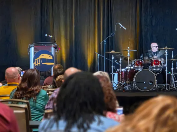 A drummer performs onstage during a NAPT ACTS 2026 conference presentation as attendees watch from the audience.