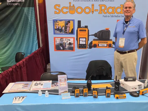 A School-Radio representative stands beside a display of push-to-talk radios and communication equipment at the NAPT ACTS 2026 trade show.