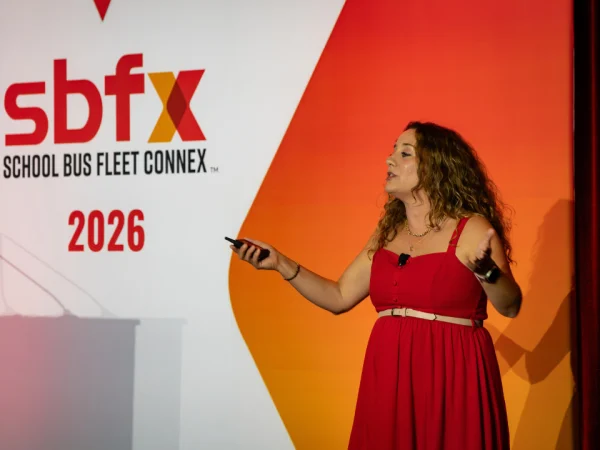 A presenter gestures while speaking onstage in front of the 2026 School Bus Fleet Connex backdrop.