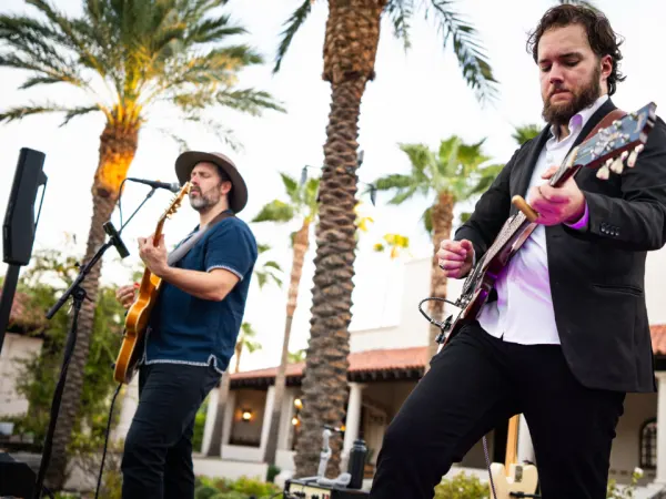 Two musicians play guitars outdoors among palm trees during an evening event at School Bus Fleet Connex.