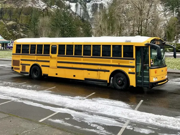 A yellow school bus is parked in front of Multnomah Falls, where water cascades down a rocky cliff surrounded by evergreen trees dusted with snow beneath a cloudy sky.