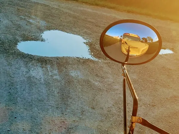 A heart-shaped puddle sits on a gravel road beside a school bus. The bus is reflected in the round crossover mirror while golden morning sunlight illuminates nearby fields.