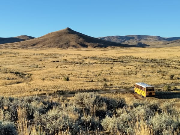 A lone yellow school bus travels along a narrow road through a vast golden grassland surrounded by rolling hills under a clear blue sky, emphasizing the scale of the landscape.