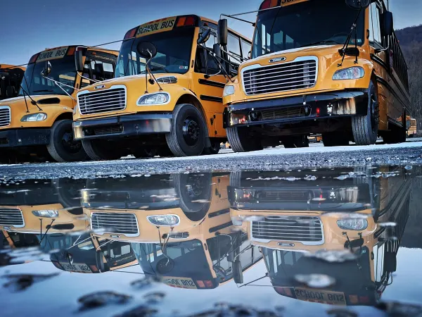 Several parked yellow school buses are reflected in a large puddle on a cold morning, creating a mirror image of the buses and blue sky across the water.