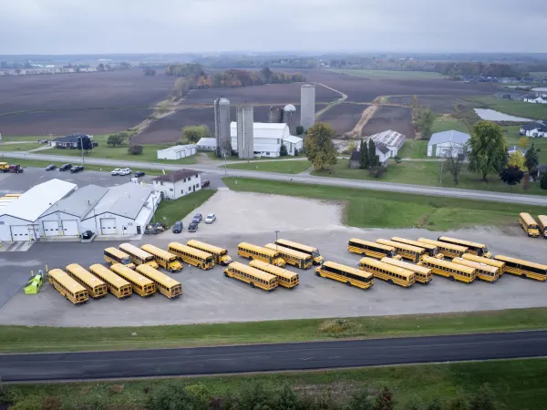 An aerial view shows dozens of yellow school buses parked around a transportation facility surrounded by farmland, silos, and rural roads under an overcast autumn sky.