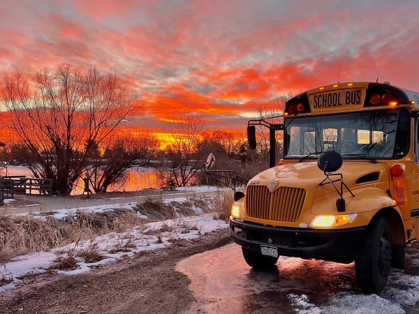 A yellow school bus is parked beside a snow-lined roadway at sunset, with vivid orange and pink clouds reflecting on a nearby pond. The bus's headlights and warning lights are illuminated against the colorful winter sky.