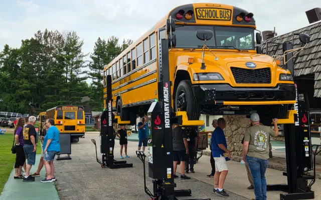 A new Blue Bird school bus is lifted on a heavy-duty Rotary vehicle hoist during an outdoor maintenance equipment demonstration as attendees gather nearby.