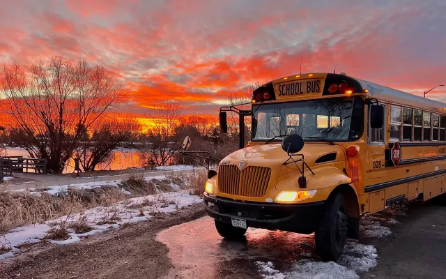 A yellow school bus is parked beside a snow-lined roadway at sunset, with vivid orange and pink clouds reflecting on a nearby pond. The bus's headlights and warning lights are illuminated against the colorful winter sky.