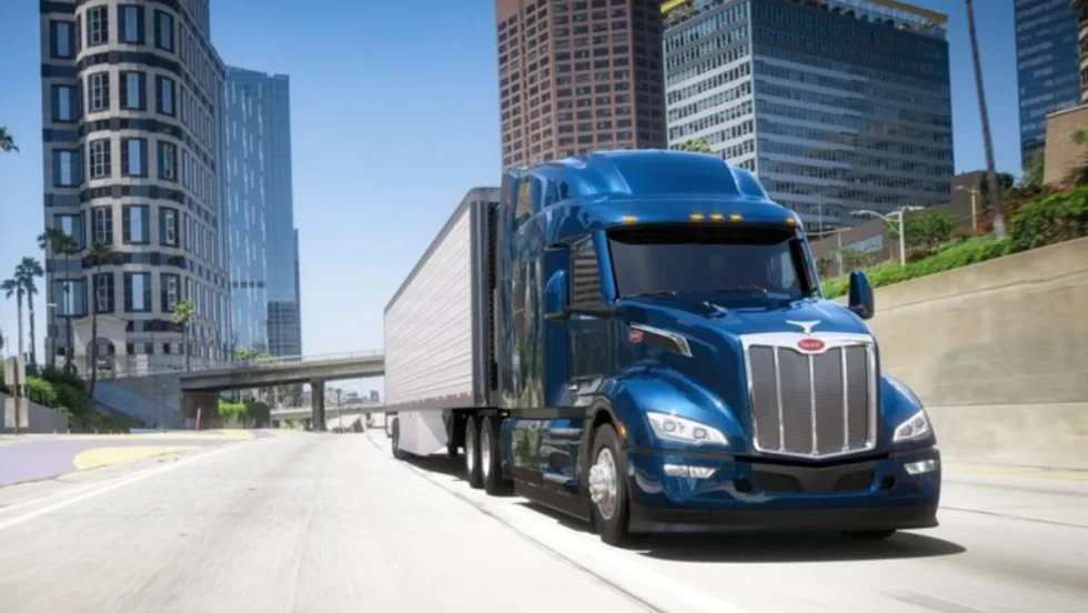 A blue Peterbilt Model 579 semi-truck driving on a city highway with tall buildings in the background.