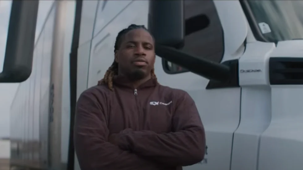 A truck driver stands with arms crossed in front of a white semi-truck, wearing a maroon jacket, featured in the ATA’s DRIVEN series highlighting professional drivers in the trucking industry.