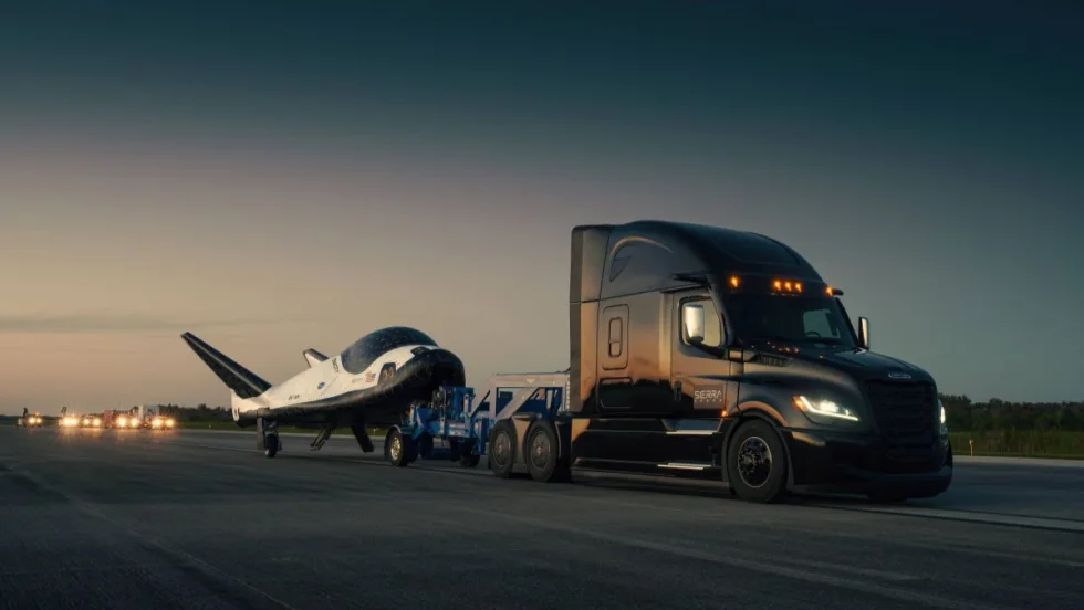 A Freightliner Cascadia semi-truck towing Sierra Space’s Dream Chaser spaceplane on a runway at dusk.