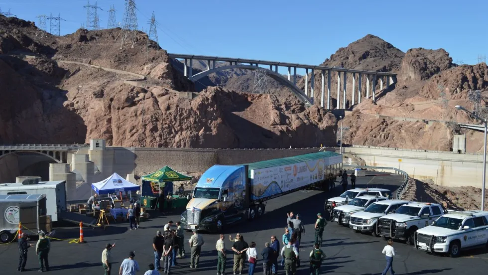 A semi-truck hauling the U.S. Capitol Christmas Tree trailer is parked near Hoover Dam, with people and law enforcement vehicles gathered nearby.