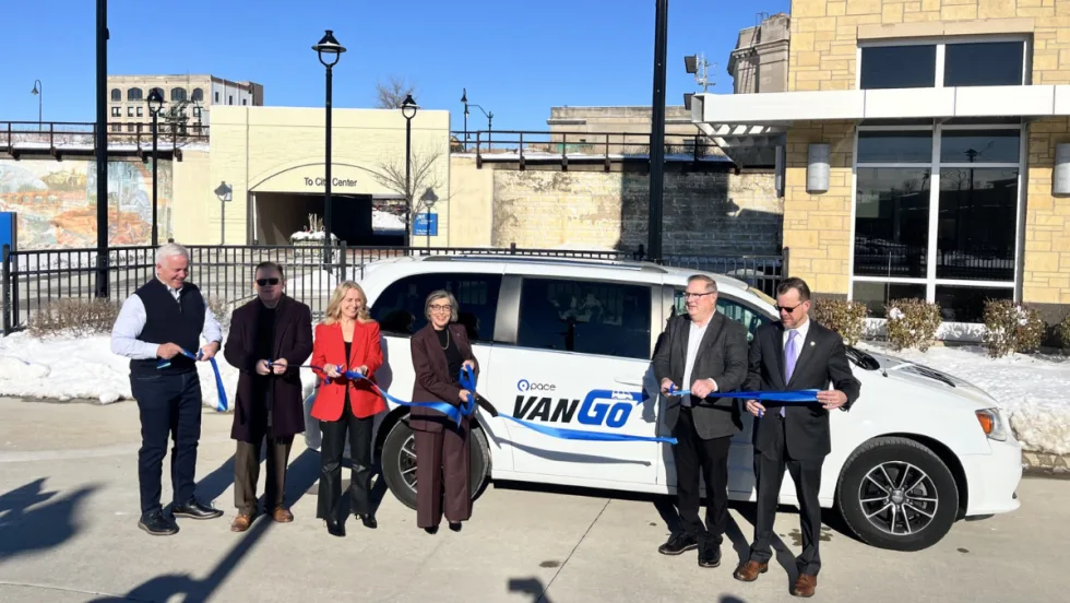 A group of officials stands in front of a white Pace VanGo minivan and holds a blue ribbon during a ribbon-cutting event outside a transit facility on a sunny winter day.