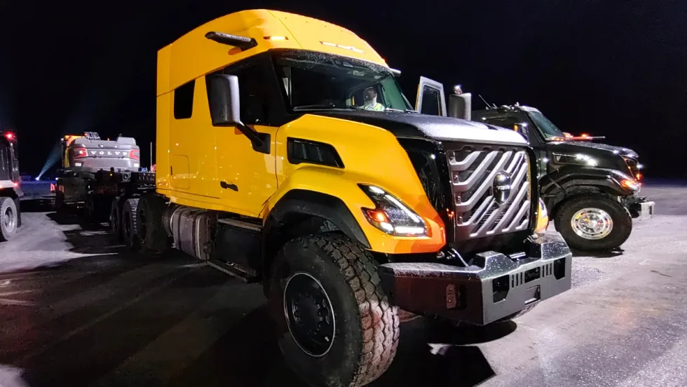 Yellow Volvo VNS heavy-haul sleeper truck with rugged tires and prominent grille, parked at night on a snow-covered lot beside a black truck, with a Volvo construction machine on a trailer behind it.