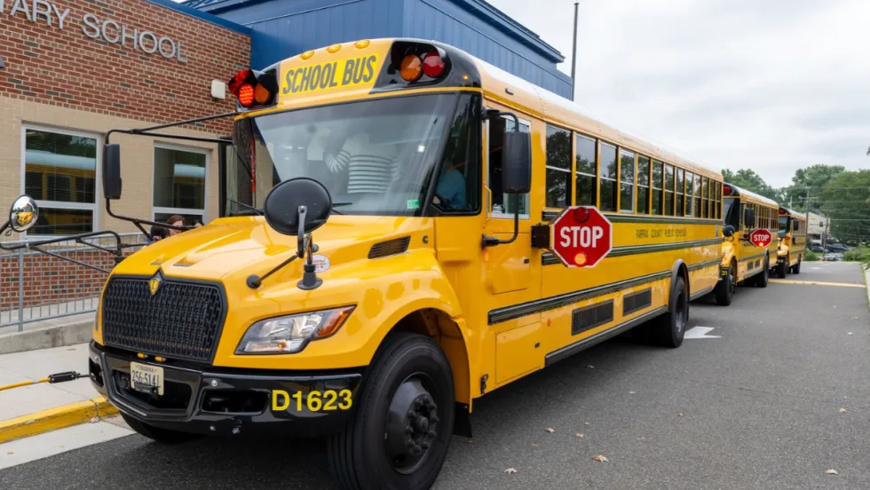 Yellow Fairfax County Public Schools buses line up outside an elementary school, with the lead bus displaying its extended stop arm and flashing red lights.