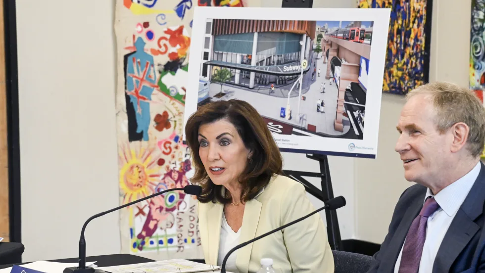 Governor Kathy Hochul sitting at a table with a microphone, Janno Lieber sits to her left.