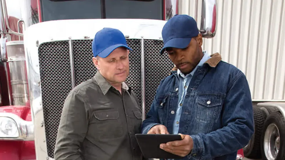 Two truck fleet managers standing in front of a semi-truck reviewing emergency response plans on a digital tablet.