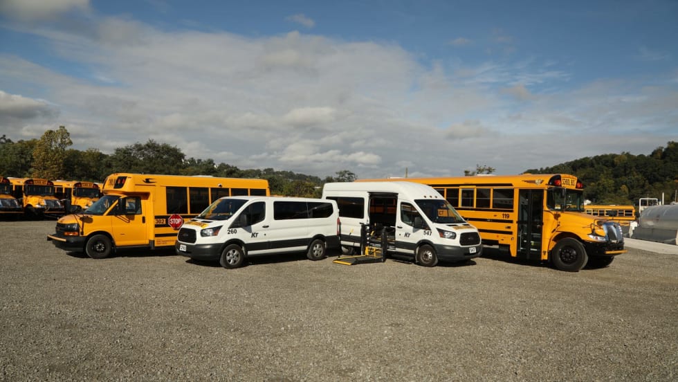 A lineup of Krise Transportation vehicles, including Type A and conventional school buses and passenger vans, with one wheelchair-accessible van shown with its lift extended.