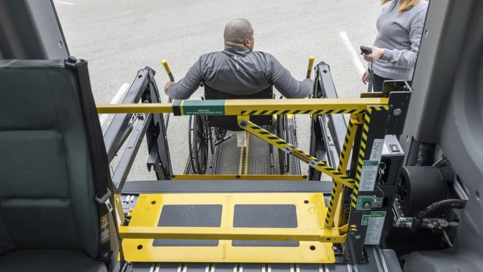 A wheelchair user operates a school bus wheelchair lift equipped with BraunAbility’s LiftSentinel barrier system as another person stands nearby with a handheld control.