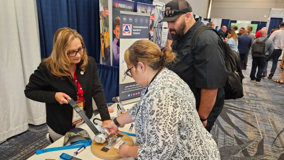 Attendees visit the AMF Bruns vendor booth in the expo hall, examining a wheelchair securement device while speaking with an exhibitor about safety equipment.