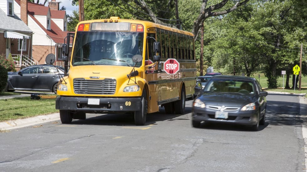  photo of a black sedan passes a stopped school bus with its sign out on a roadway